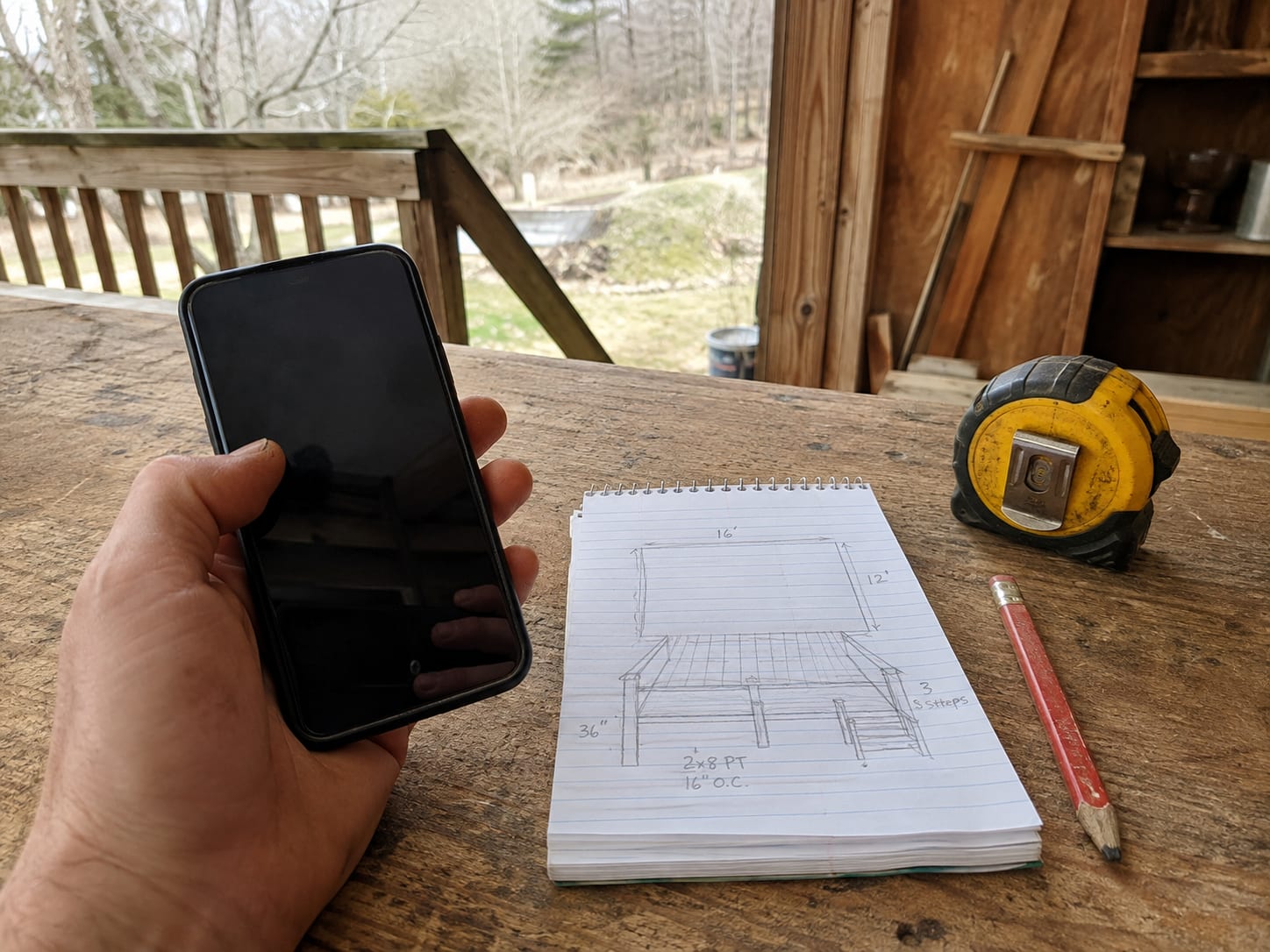 A phone held over a workbench beside a notepad sketch of a deck marked up with dimensions — 16 by 12 feet, 36 inch rail, 2x8 pressure-treated joists at 16 inches on centre — with a tape measure and carpenter's pencil alongside