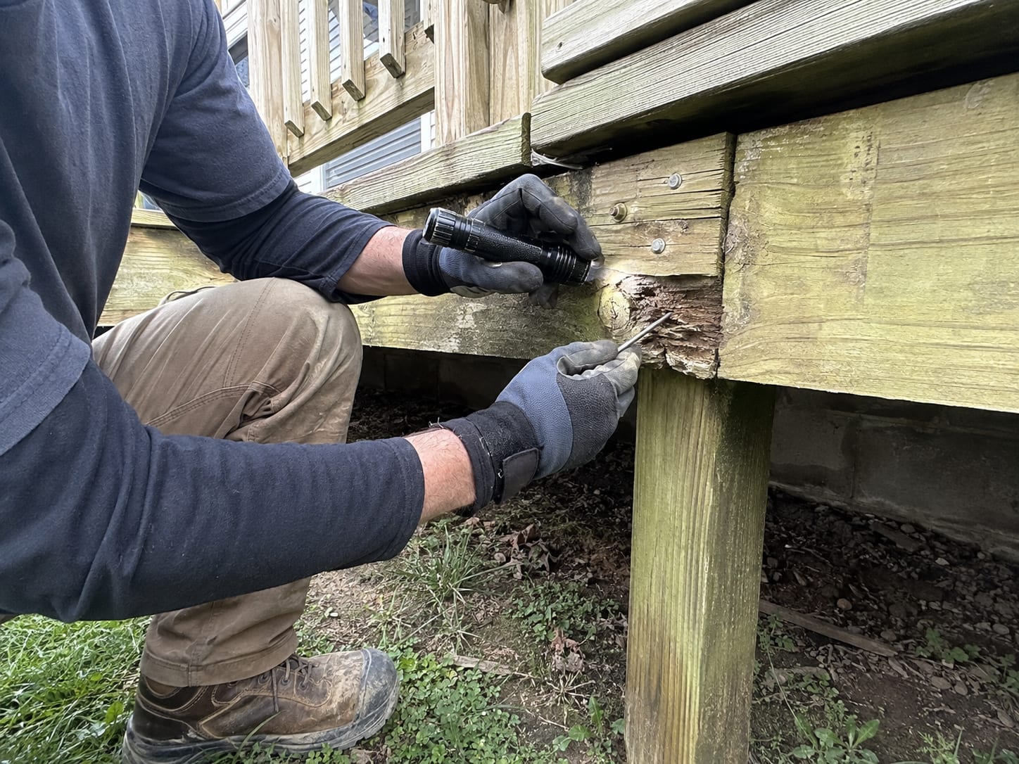 Kneeling under a deck to inspect the framing, using a flashlight and a probe to test a rotted band board where it meets a support post