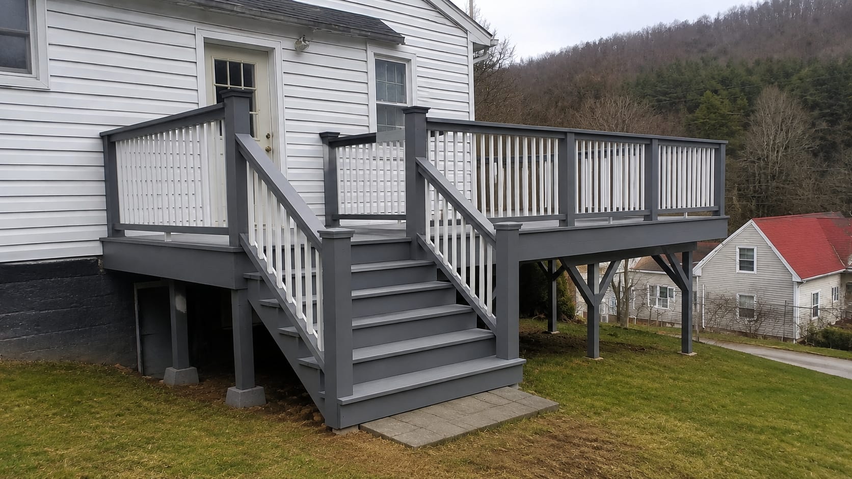 Raised wood deck built by Mackwood Construction on a white clapboard West Virginia home — grey deck boards and posts with white railing balusters, wide steps down to a paver pad, wooded hillside behind