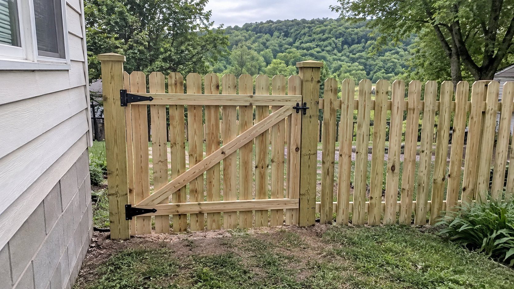 New pressure-treated wood gate with black strap hinges hung in a picket fence beside a house