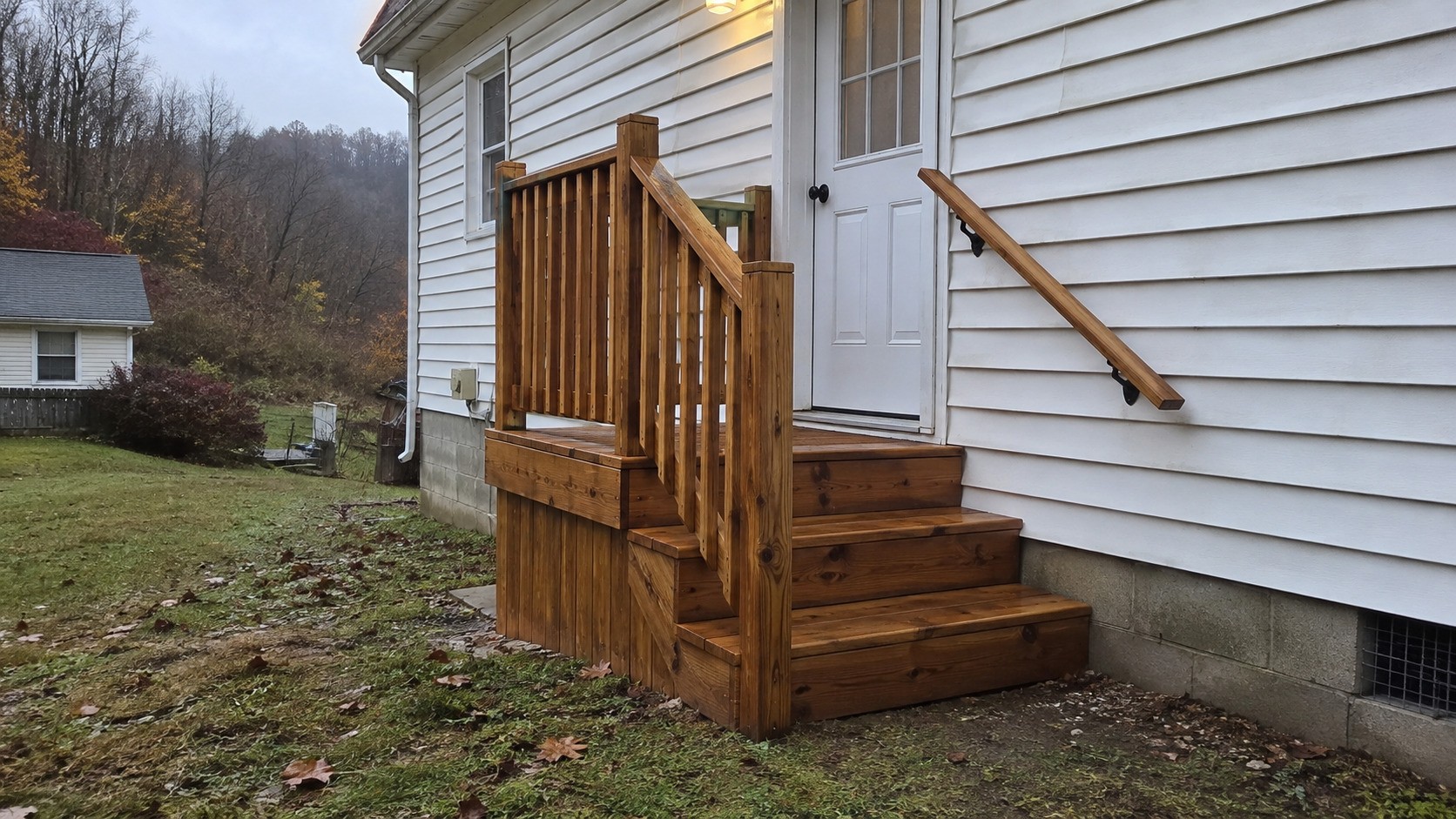 New stained wood back steps with a railing and a wall-mounted handrail beside the door of a sided house