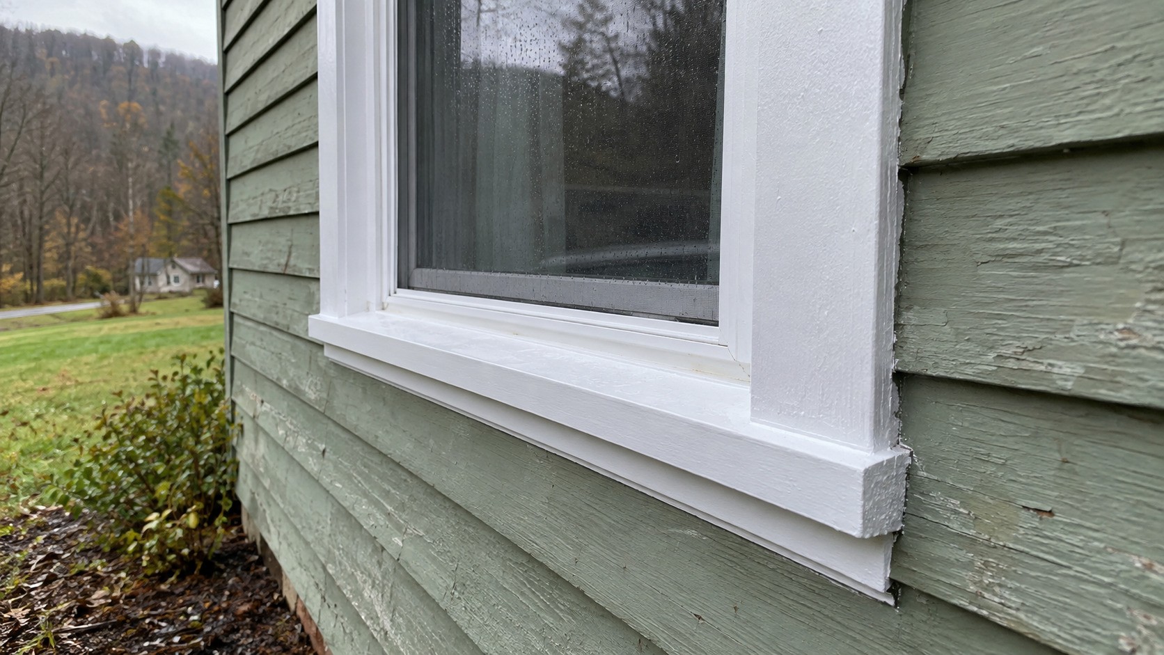 New white window trim and sill fitted against weathered green wood siding on an older house